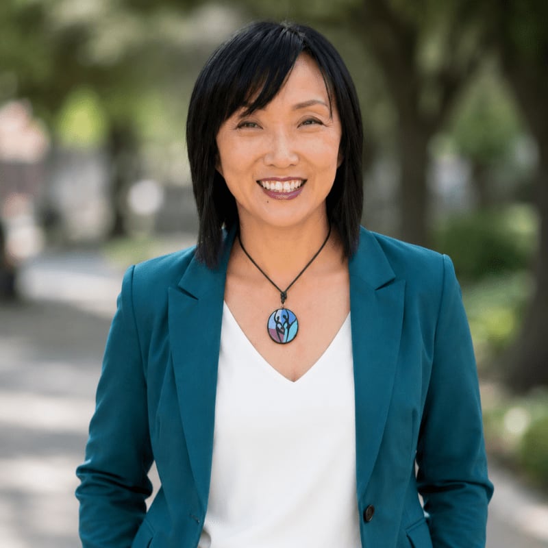A smiling woman in a teal blazer and white top stands in the foreground with a blurred background of trees and a walkway.