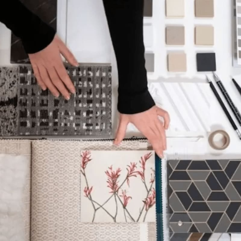 Hands arrange fabric swatches and color samples on a white surface, with pencils and tape in the background.