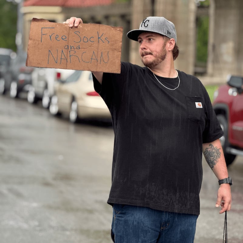A man in a black t-shirt and jeans holds a cardboard sign that reads "Free Socks and NARCAN" while standing on a wet street with cars and a building in the background.