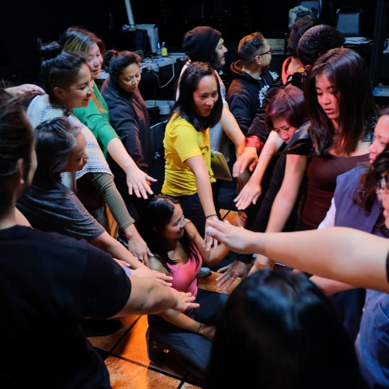 A group of people with their hands in the center of a circle are gathered in a dimly lit room with stage equipment in the background.