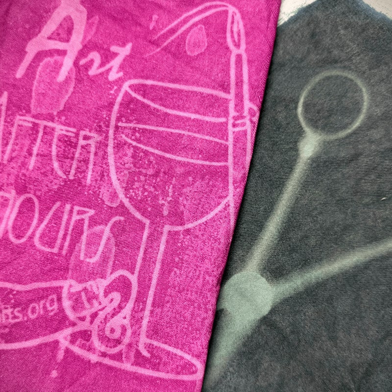 A bright pink fabric with "Art After Hours" and a wine glass printed on it is placed next to a dark gray fabric with a faint outline of a caliper.