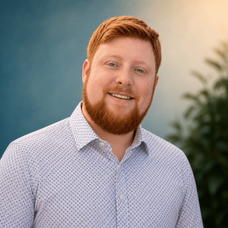 A smiling man with red hair and a beard in a patterned shirt stands against a blurred background of blue sky and green foliage.