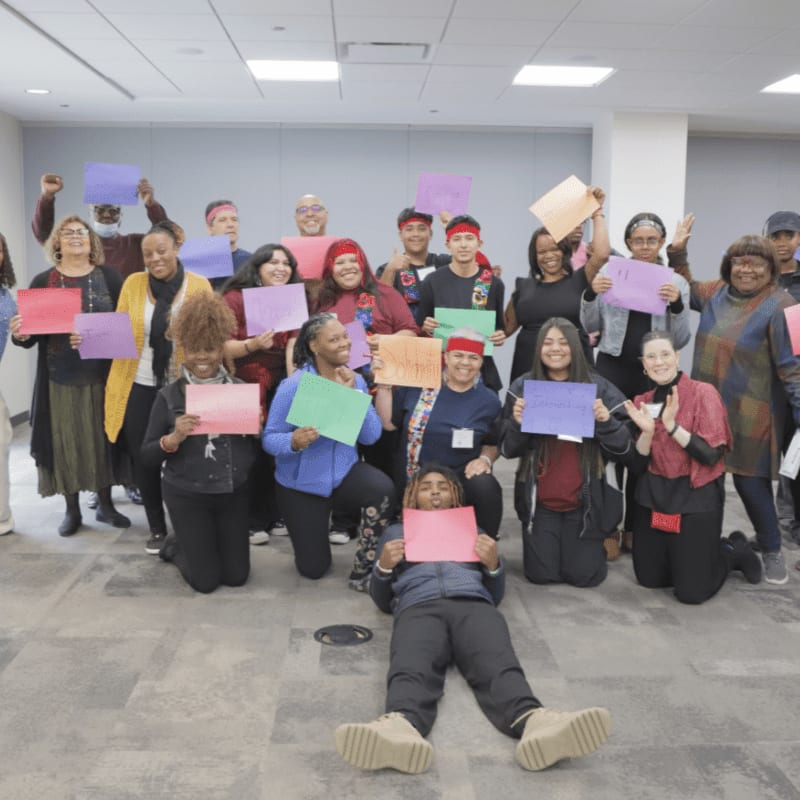 A diverse group of people in the foreground holds colorful signs, while the background shows a brightly lit room with a plain wall.