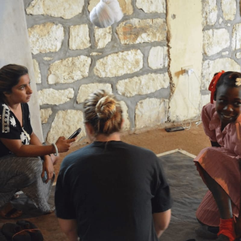 In the foreground, a woman interviews a young girl while another woman faces away, and the background features a stone wall and a doorway.