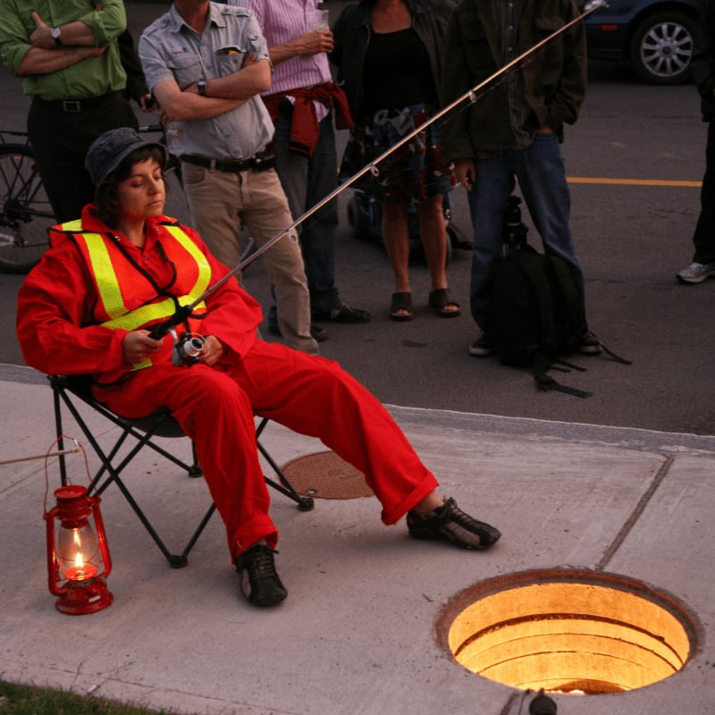 A woman in a red jumpsuit and safety vest fishes with a rod over a manhole while a crowd watches in the background.