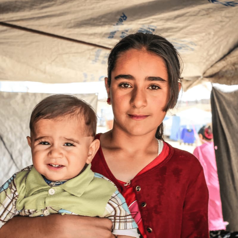 A young girl cradles a baby in her arms, with a tent and blurred figures in the background.