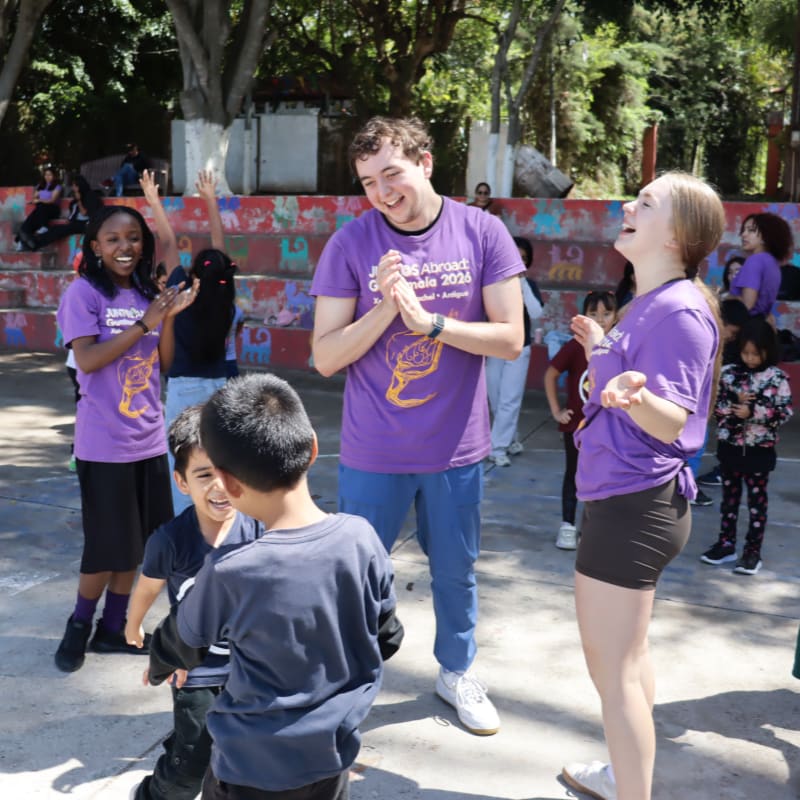 In the foreground, children in dark shirts laugh and play, while in the background, smiling young adults in purple shirts clap and cheer them on amidst a colorful, outdoor setting.