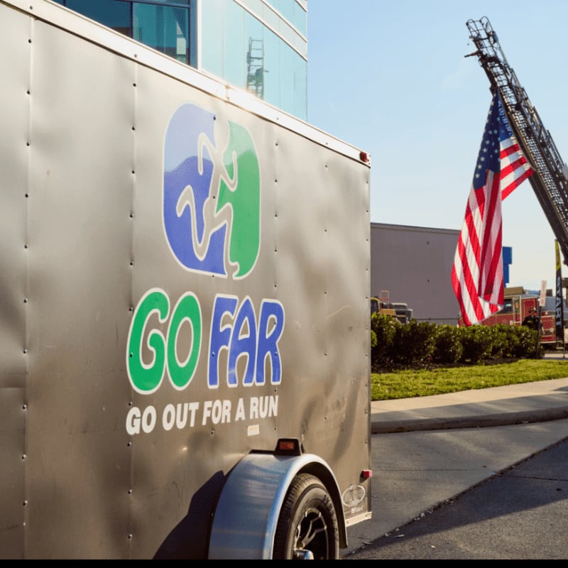 A dark trailer with a "GO FAR" logo is parked in front of a building, with a fire truck ladder and American flag in the background.