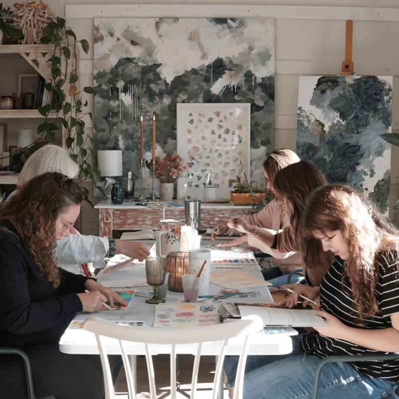 Several women are gathered around a table in a bright art studio, working on their paintings with large abstract art pieces hanging on the wall behind them.
