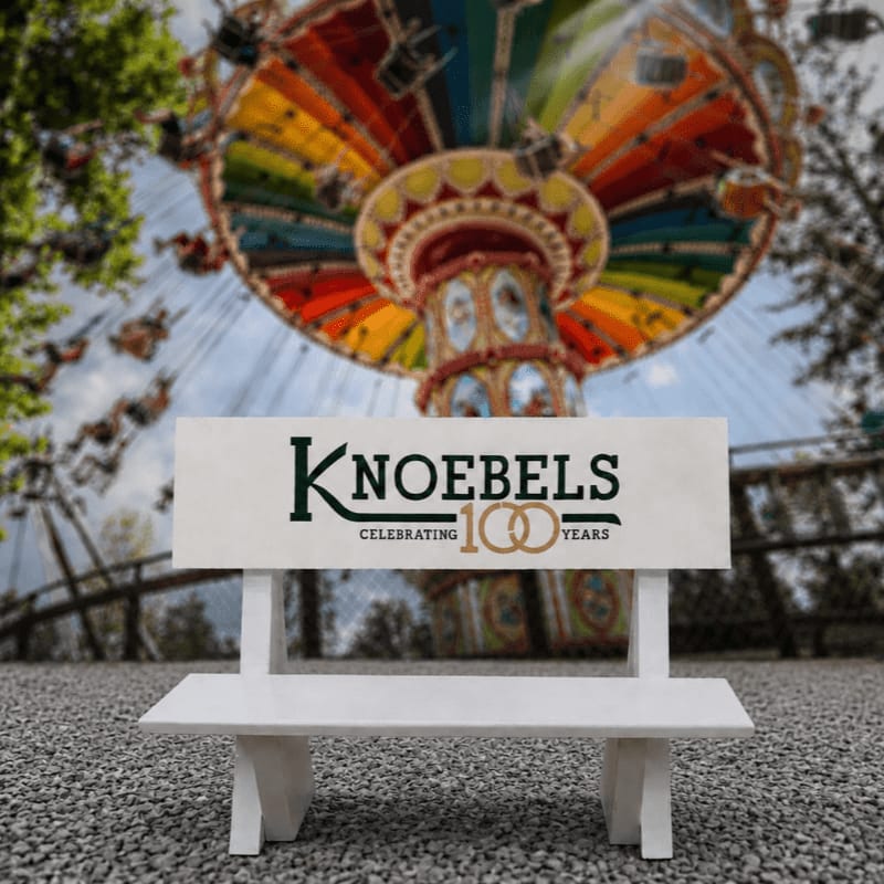 A white bench with the Knoebels 100 Years logo sits in the foreground, with a colorful amusement park ride blurred in the background.