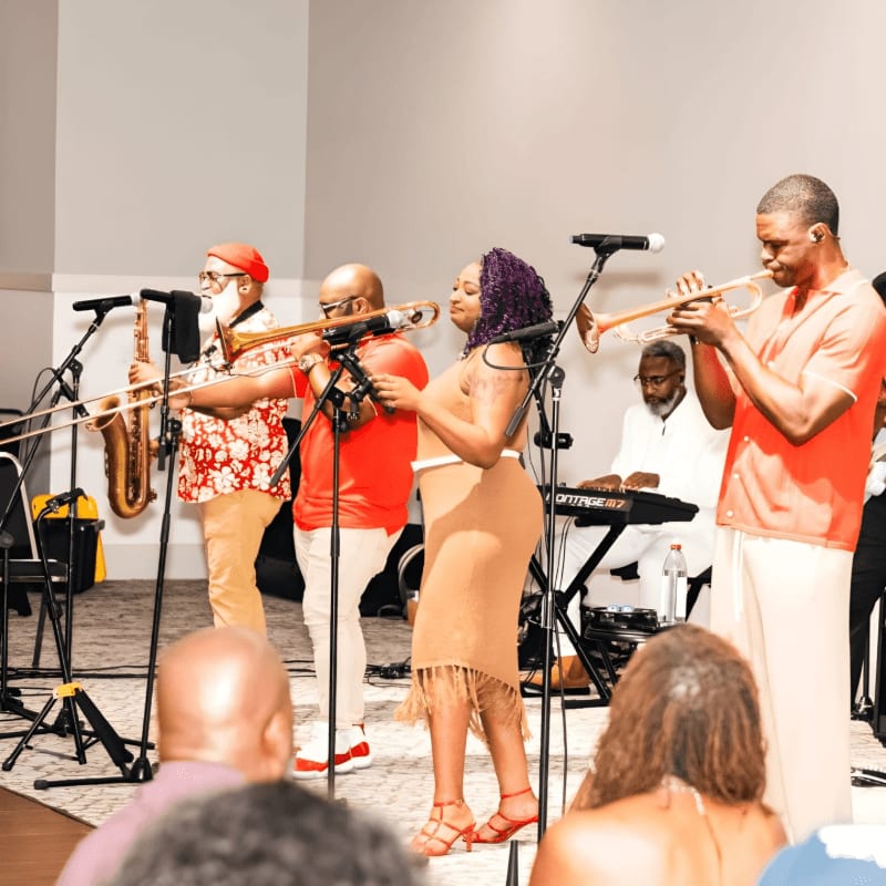 A band plays jazz music on a stage with a keyboardist in the background, while an audience watches from the foreground.