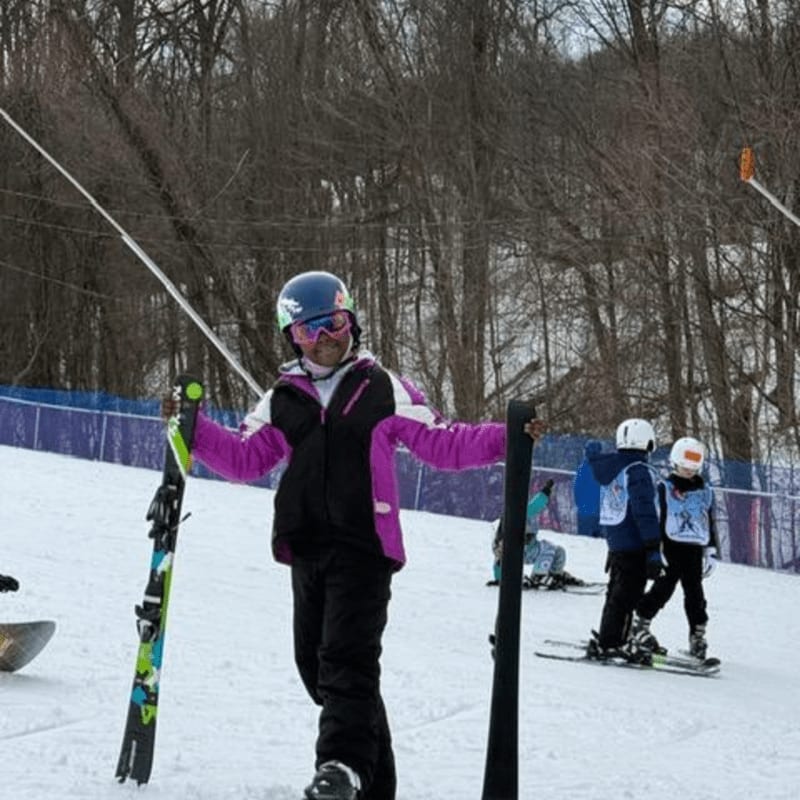 A skier in a purple and black jacket stands in the snowy foreground holding skis, with bare trees and a ski lift visible in the background.