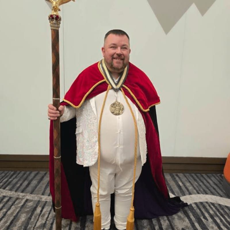 A man in ceremonial robes and a jeweled staff stands against a patterned wall.