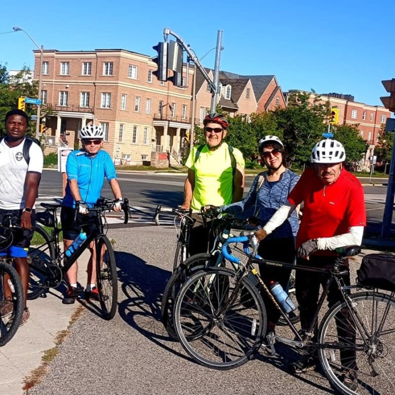 Five cyclists stand with their bikes on a paved path in the foreground, with suburban houses and streetlights visible in the background under a clear blue sky.