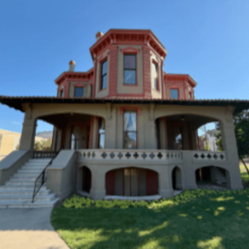 A grand Victorian mansion with a wide porch and manicured lawn stands against a clear blue sky.