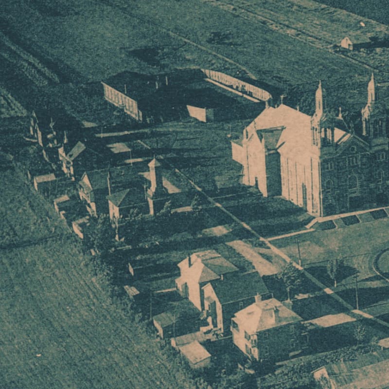 A large, ornate church with a steeple dominates the right side of the frame, while a row of houses and a long, low building stretch across the left, all set against a backdrop of fields and shadows.