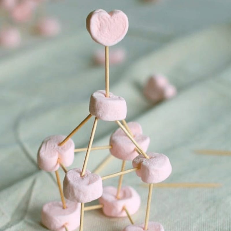 A delicate marshmallow tower, topped with a heart-shaped candy, stands on a soft green surface with blurred marshmallows scattered in the background.