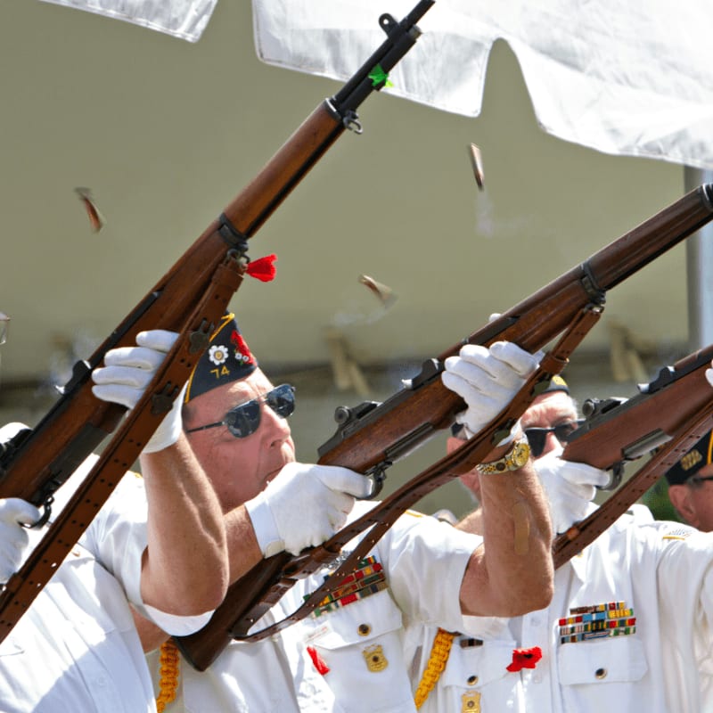 Veterans fire rifles in a ceremonial salute outdoors.