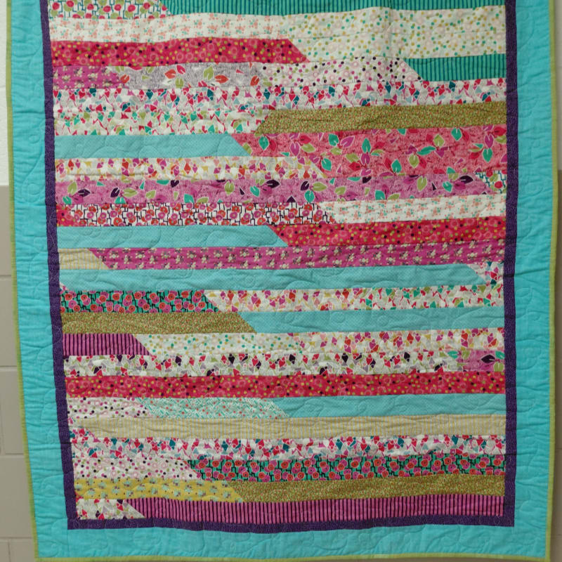 A colorful, handmade quilt with a striped pattern of floral and geometric fabrics is displayed against a plain, light-colored wall.