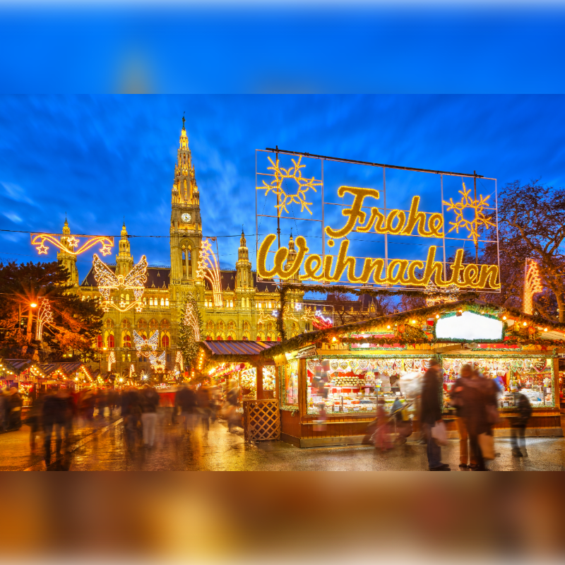 A bustling Christmas market with illuminated stalls and people in the foreground is set against the backdrop of a grand, ornate building adorned with festive lights and a "Frohe Weihnachten" sign.