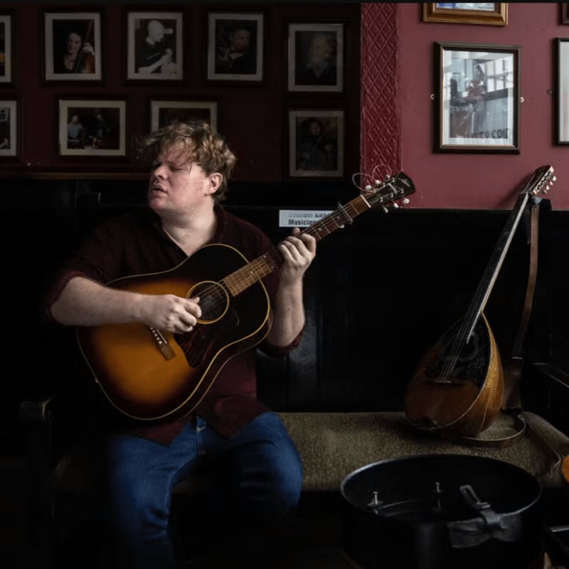 A man plays an acoustic guitar in the foreground, while framed portraits adorn the wall in the background.