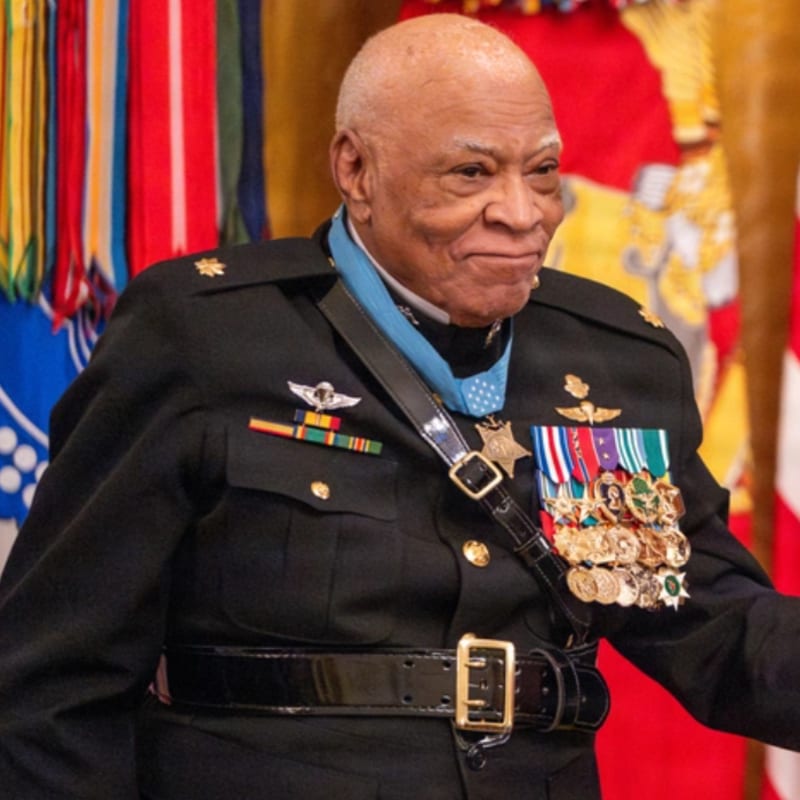A distinguished elderly man in a military uniform adorned with numerous medals stands before a backdrop of colorful flags and banners.