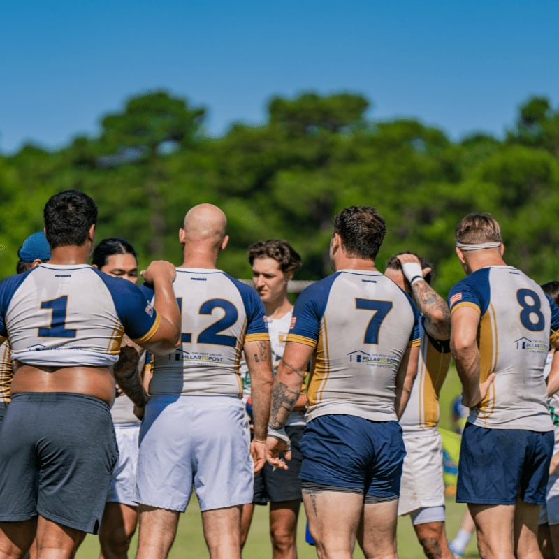 A group of rugby players in matching uniforms stand on a grassy field with trees in the background under a clear blue sky.