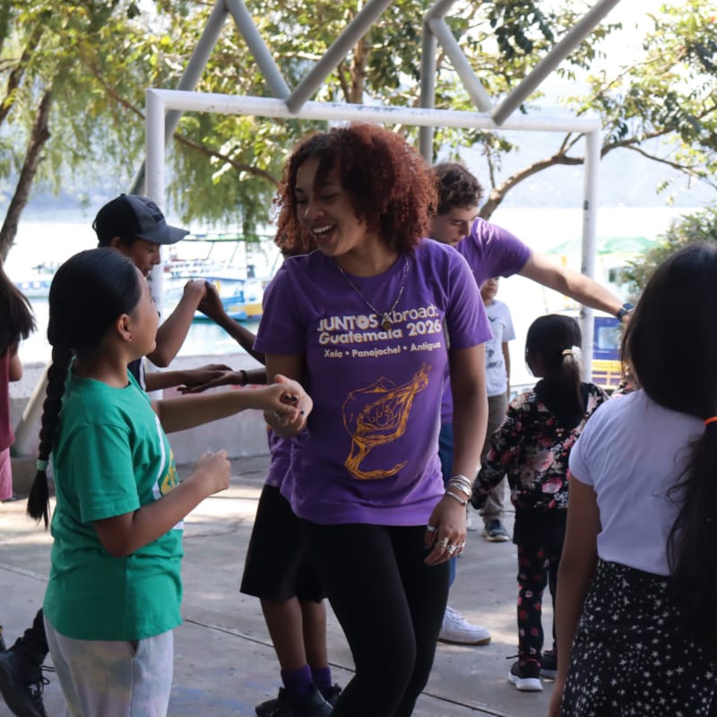 A woman in a purple shirt laughs with children in a courtyard, with trees and a body of water visible in the background.