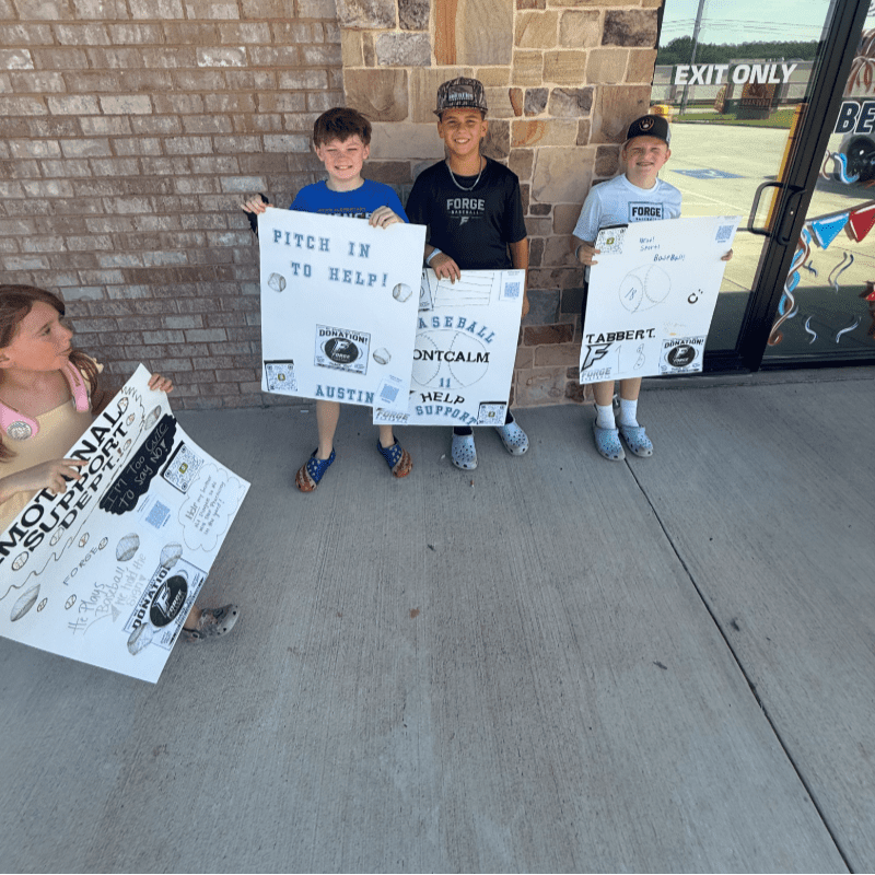 Four children stand on a sidewalk holding signs, with a brick building and an exit sign in the background.