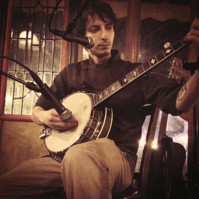 A man plays a banjo in the foreground, with a dimly lit, windowed background.