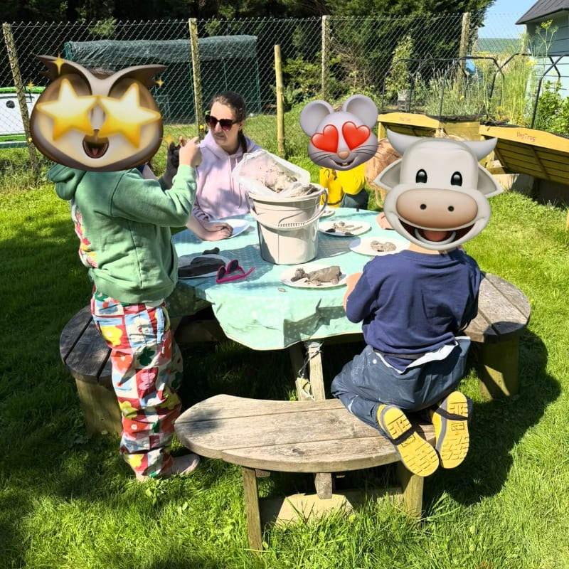 Four children with emoji masks sit around a picnic table in a grassy yard, with a fence and trees in the background.