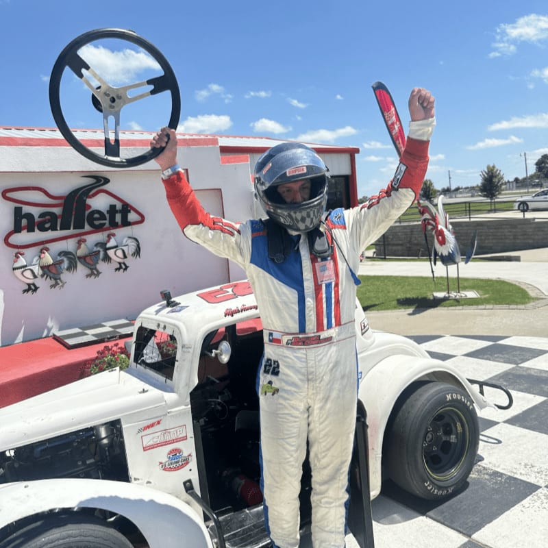 A race car driver in a white and red suit stands triumphantly with a steering wheel raised in the air in front of a building with a "hallett" sign and a checkered ground, while a blue sky with clouds forms the background.