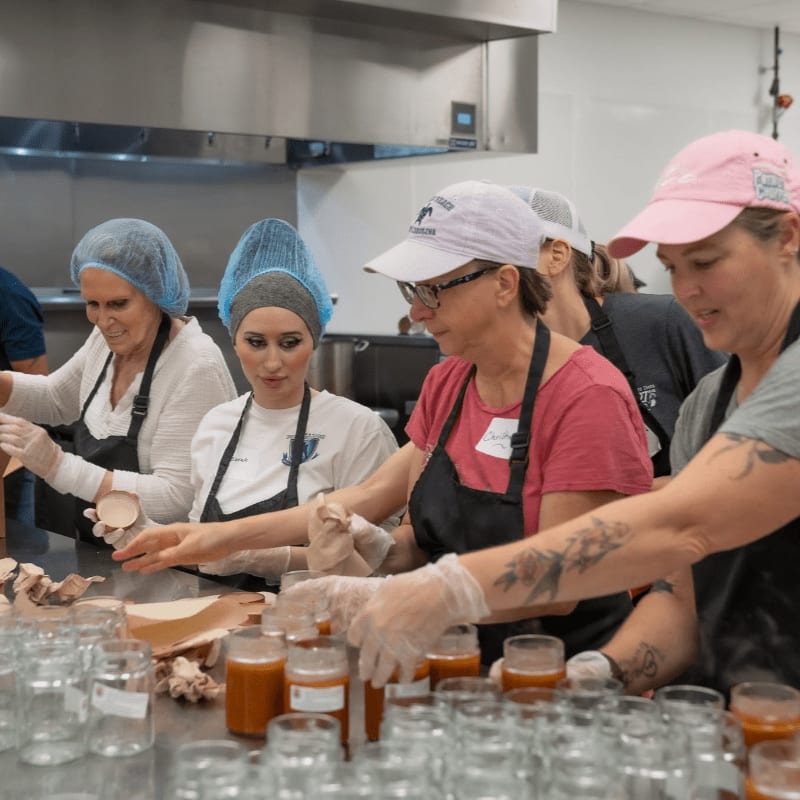 In the foreground, several women in aprons and hairnets are filling jars with food, while the background shows a commercial kitchen setting.