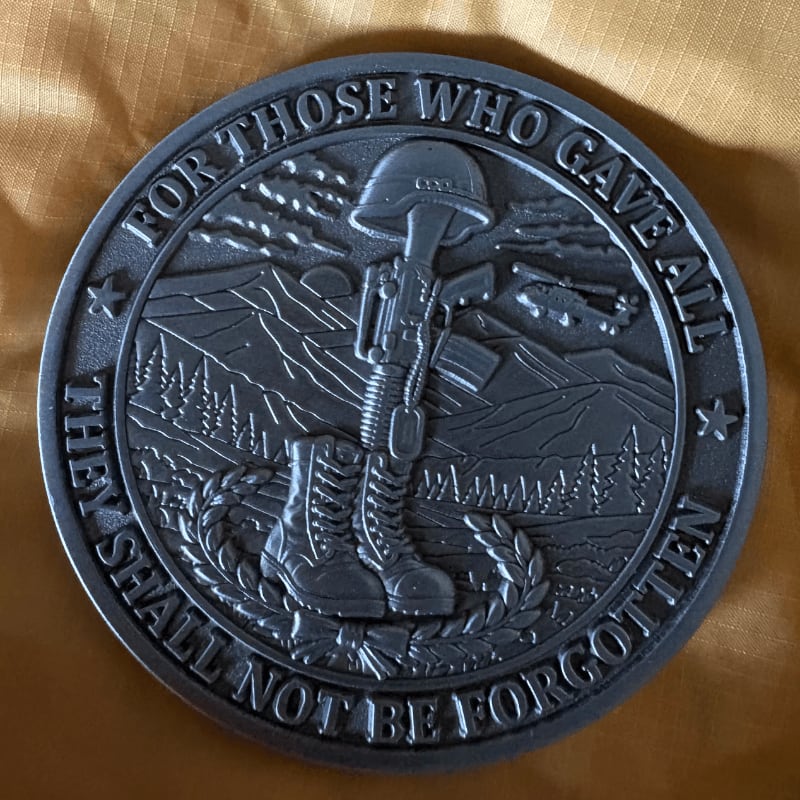 A memorial coin with a fallen soldier's helmet and rifle in the foreground rests on a textured, golden-brown surface, while a mountainous landscape with a helicopter is etched into the background.