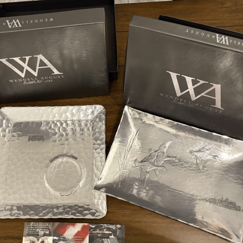 Two decorative metal trays, one with a hammered finish and the other with a duck etching, sit in front of their respective Wendell August gift boxes.