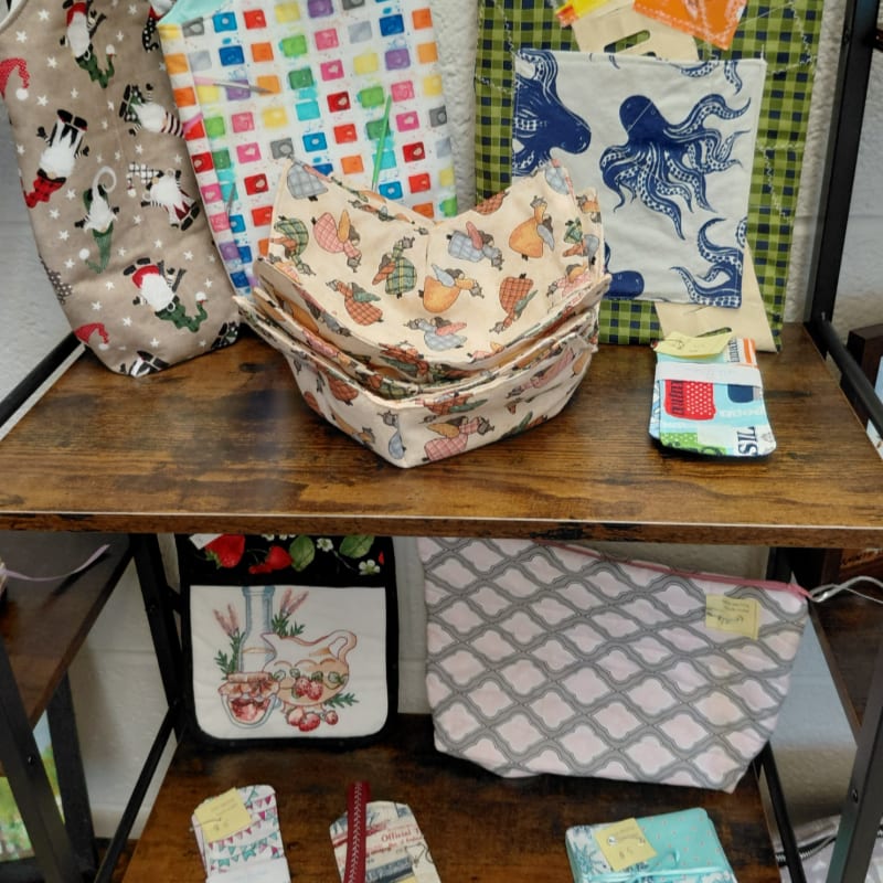 Colorful fabric bowls and bags are displayed on wooden shelves, with a patterned bag and a green checkered fabric in the background.