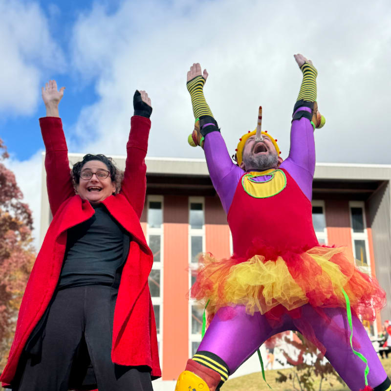 Two people in colorful costumes with arms raised cheer in front of a building under a cloudy sky.