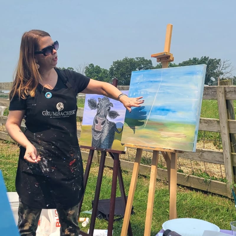A woman in an apron points to a landscape painting on an easel, with a cow painting and a wooden fence visible in the background.