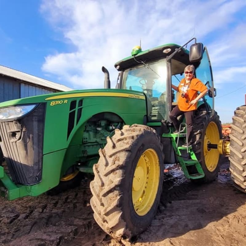 A woman stands on the steps of a large green tractor in the foreground, with a barn and a bright blue sky in the background.