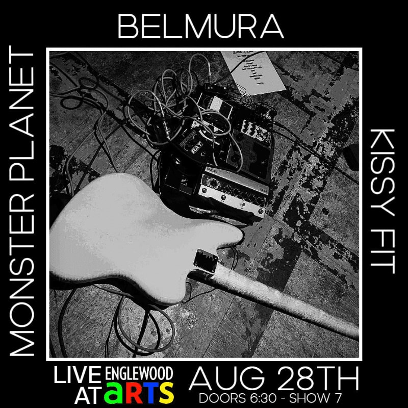 A white electric guitar and audio equipment rest on a wooden floor in the foreground, with band names and event details filling the background.