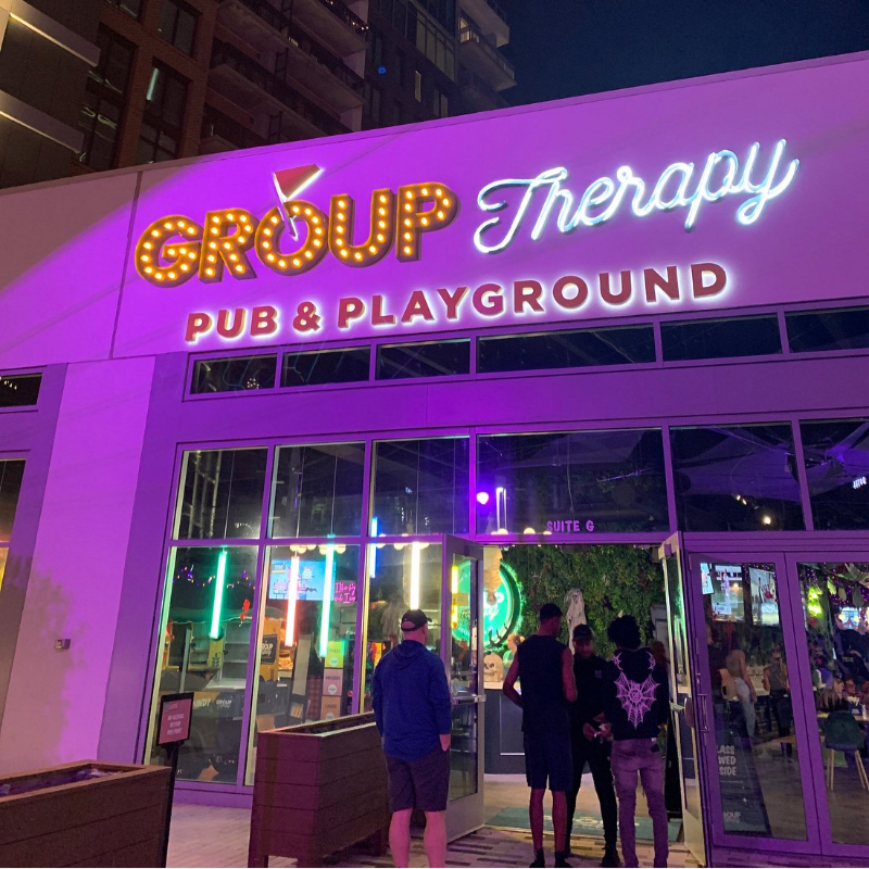 A group of people stand outside a brightly lit "Group Therapy Pub & Playground" with neon signs and a modern building in the background.