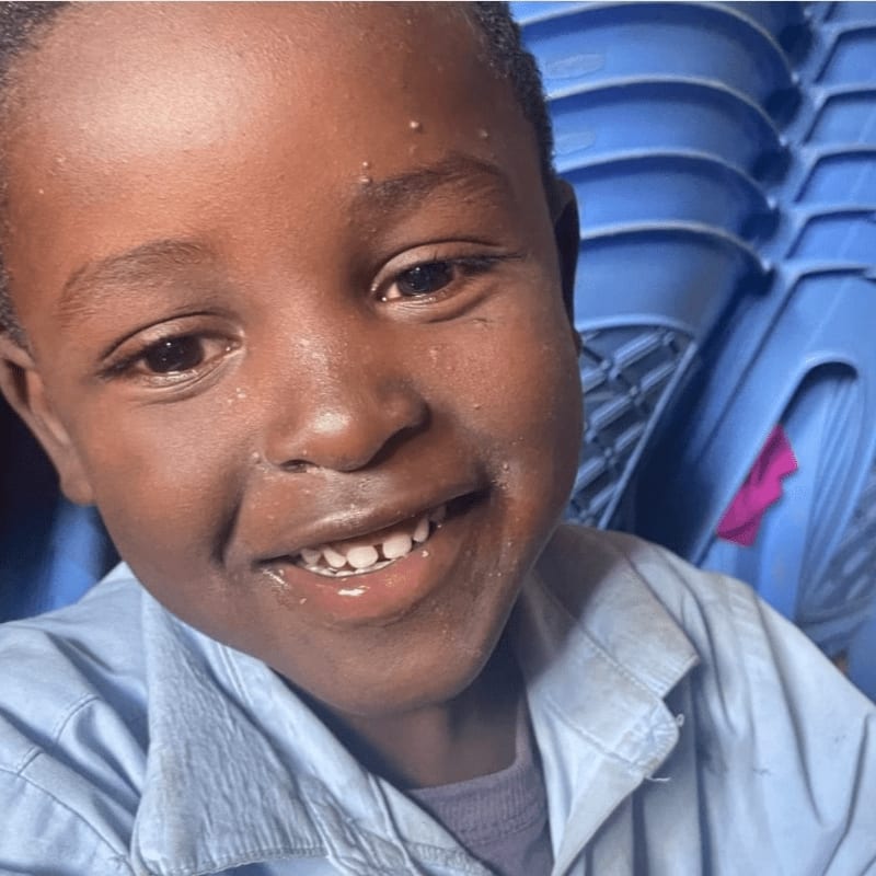 A young boy with a bright smile is in the foreground, with a stack of blue chairs in the background.
