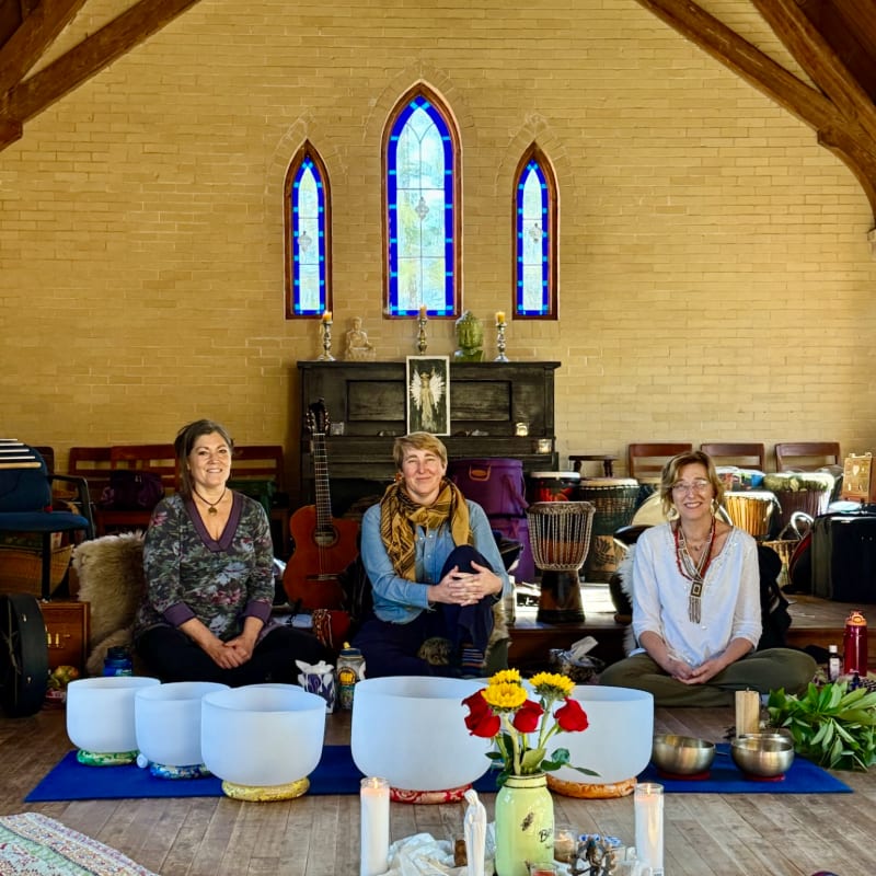 Three women sit cross-legged in front of large crystal singing bowls, with a backdrop of drums, instruments, and stained-glass windows in a rustic room.