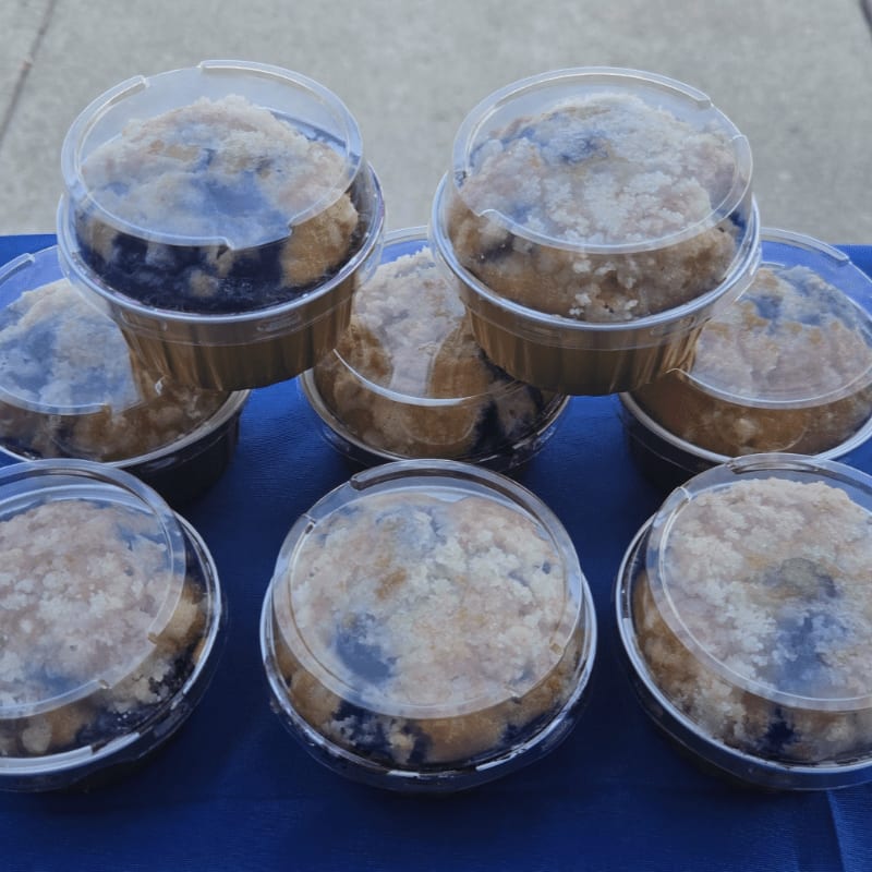 Several blueberry muffins in clear plastic containers are arranged on a blue surface with a blurred outdoor background.