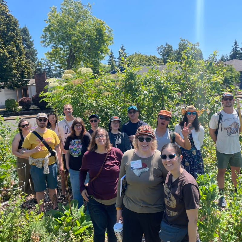 A group of people stand in a lush garden with houses and trees visible in the background.