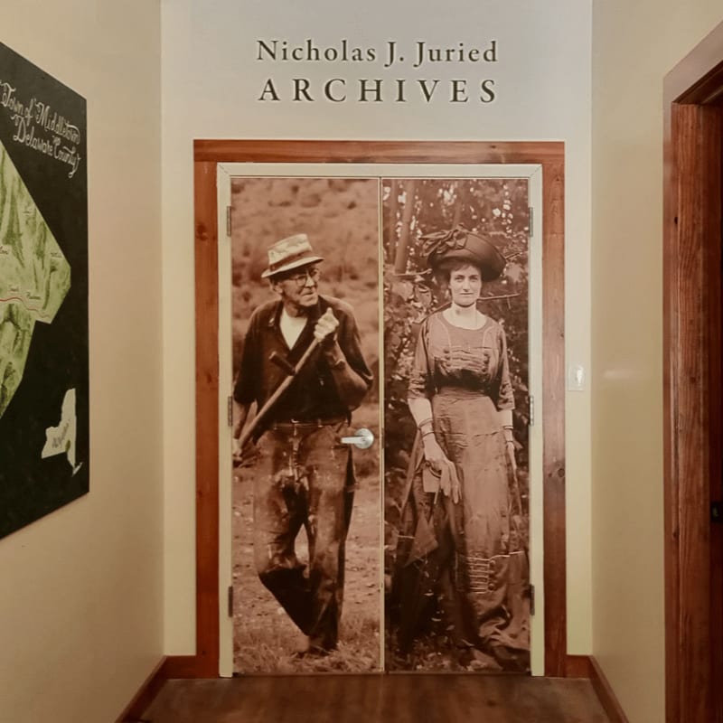 A hallway features double doors adorned with sepia-toned portraits of a man and a woman, with the "Nicholas J. Juried ARCHIVES" sign above and a map on the left wall.