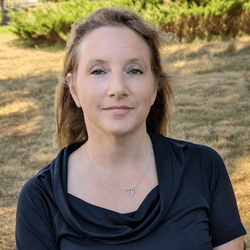 A woman with light brown hair and a dark blue cowl-neck shirt is in the foreground, with a blurred background of grass and bushes.