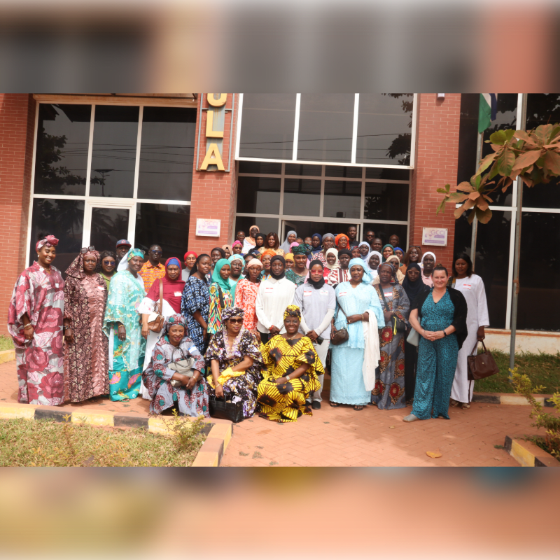 A large group of women in colorful attire pose for a photo in front of a brick building with large windows.
