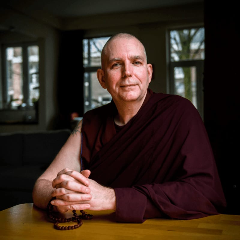 A bald man in maroon robes sits with his hands clasped on a wooden table, a mala resting beside them, while a dimly lit room with windows is visible in the background.
