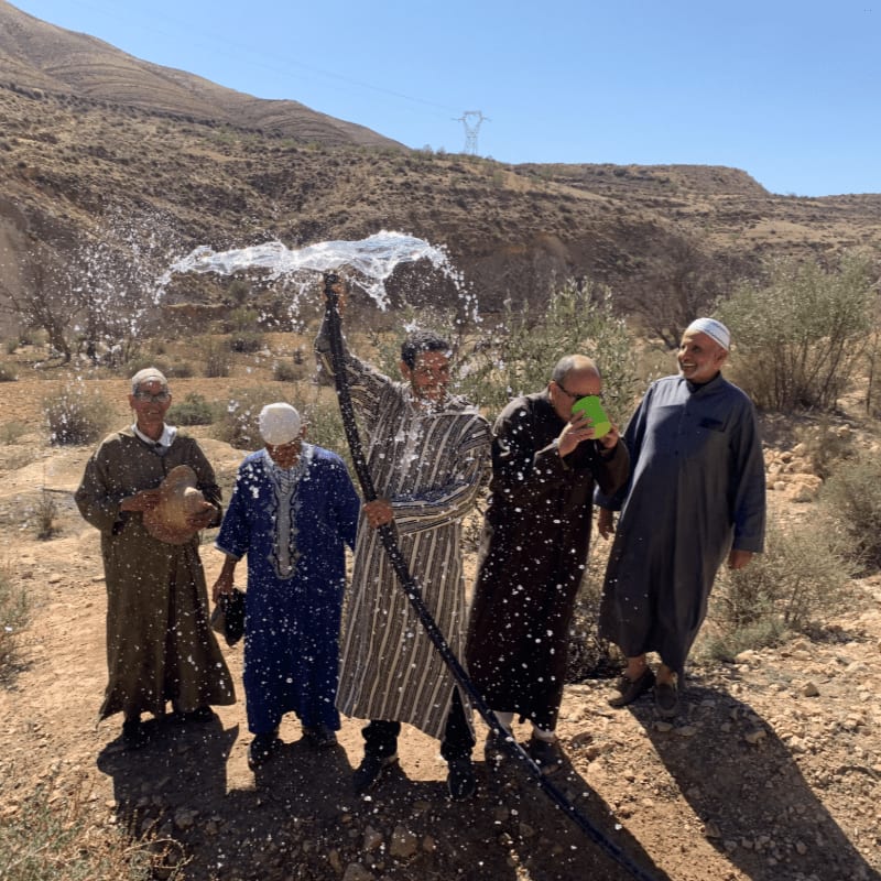 Five men in traditional clothing stand in a dry, rocky landscape as one sprays water from a hose, with arid hills and a clear blue sky in the background.
